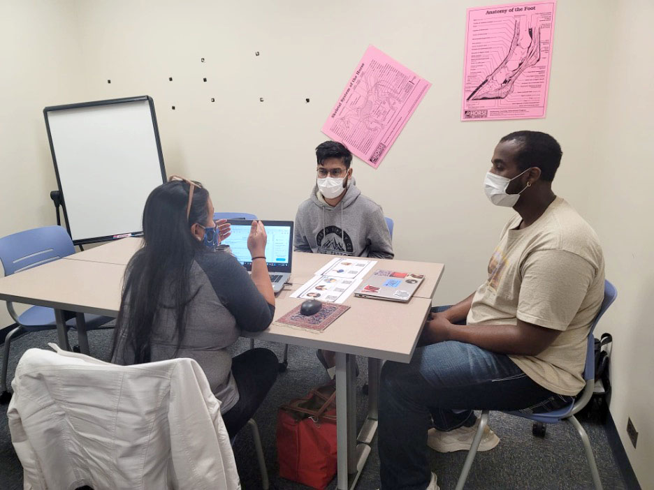 Three people discuss at a table with a laptop and materials while seated in a classroom setting.