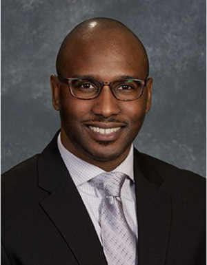 Smiling man in a suit and tie, wearing glasses, against a textured gray background.